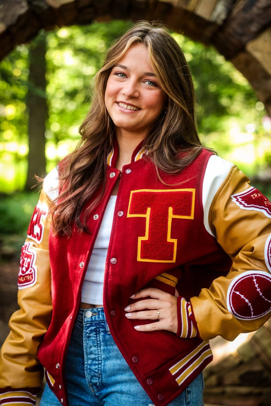 Senior portrait of a young woman in a red and gold varsity jacket beneath a stone arch